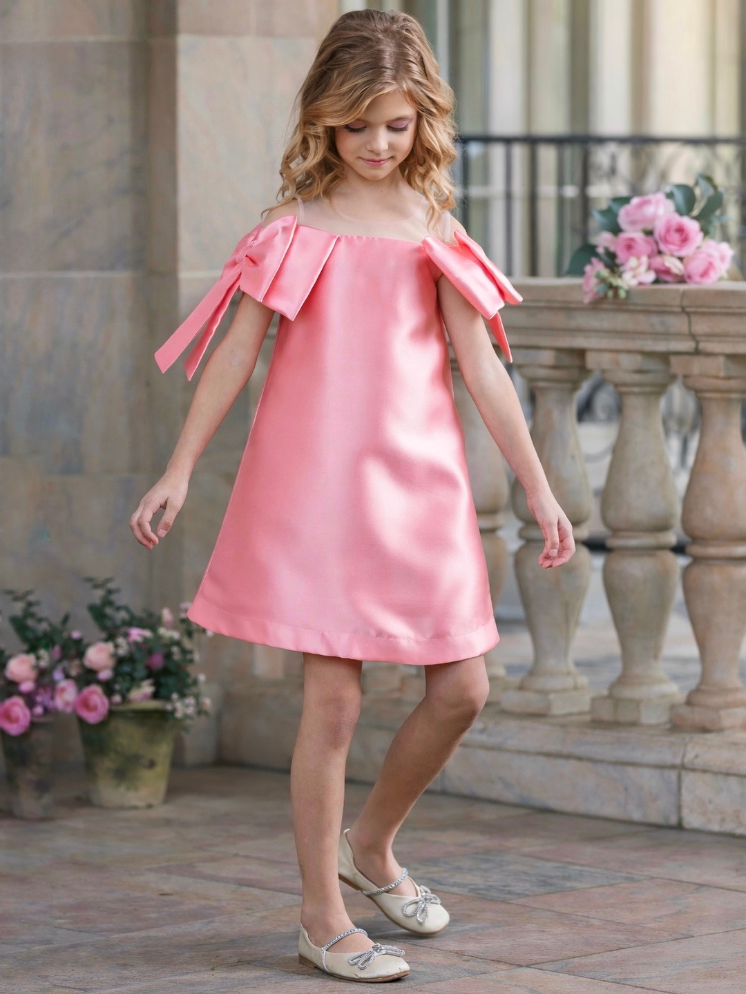 Girl wearing a blush pink satin shift dress with oversized shoulder bows, perfect for graduation, Easter, church, and spring pictures