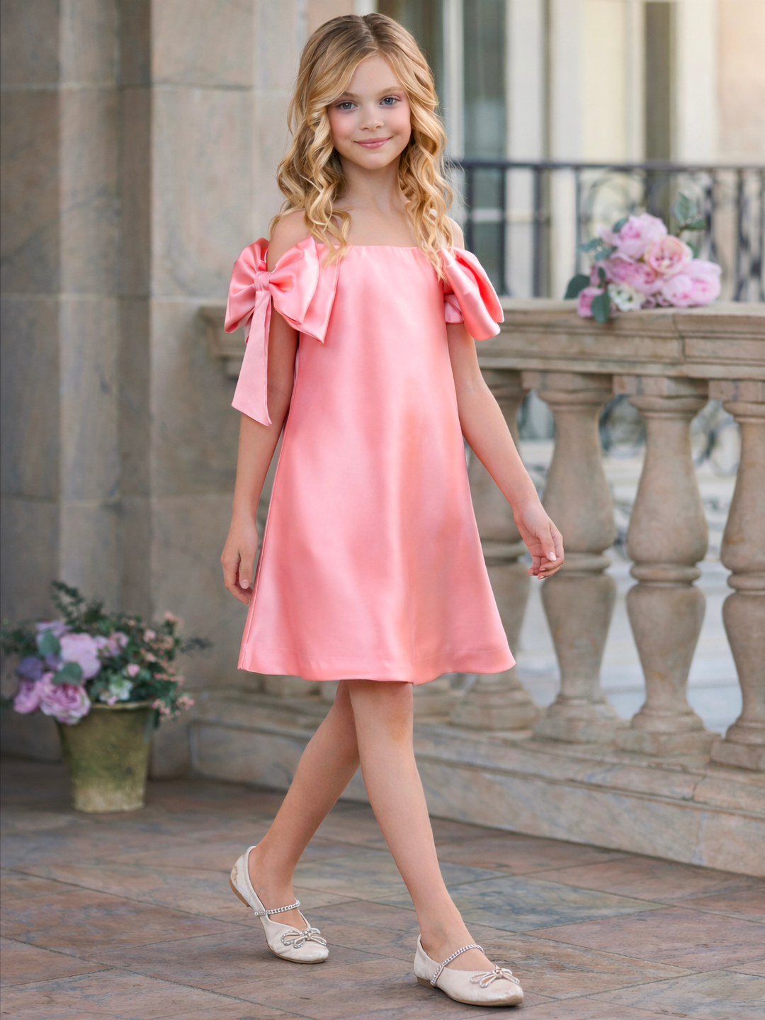 Girl wearing a blush pink satin shift dress with oversized shoulder bows, perfect for graduation, Easter, church, and spring pictures