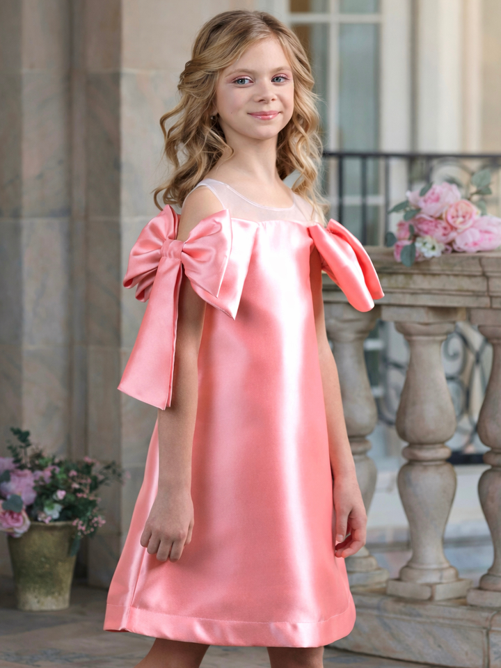 Girl wearing a blush pink satin shift dress with oversized shoulder bows, perfect for graduation, Easter, church, and spring pictures