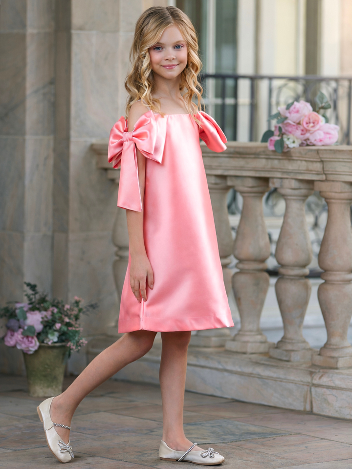 Girl wearing a blush pink satin shift dress with oversized shoulder bows, perfect for graduation, Easter, church, and spring pictures