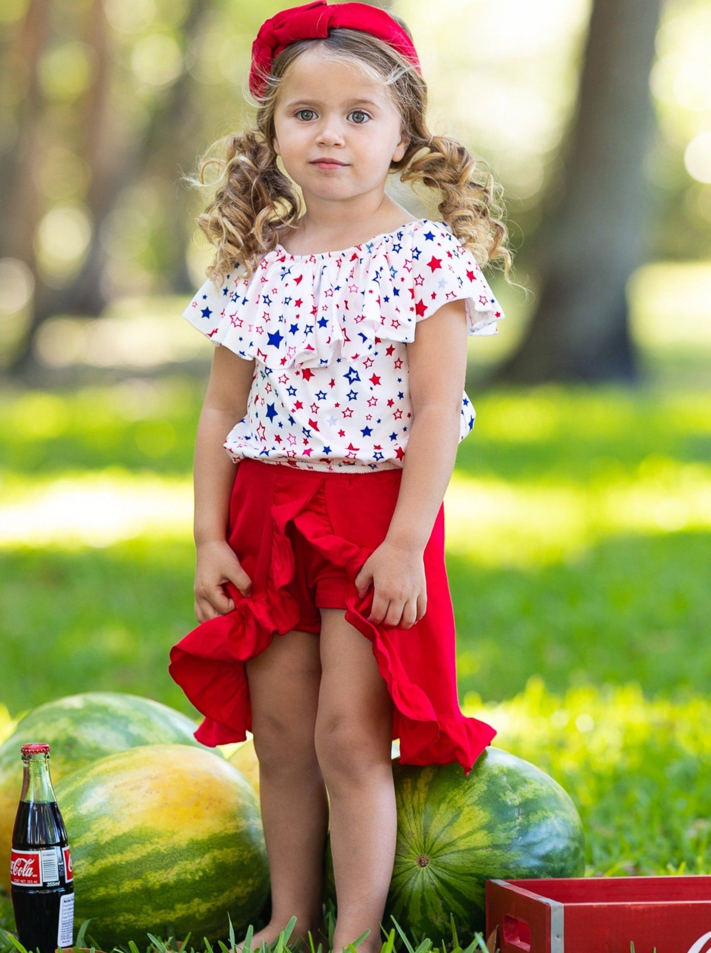Girls three piece set features a white ruffle top with red and blue star print and red shorts with matching overlay wrap ruffle skirt