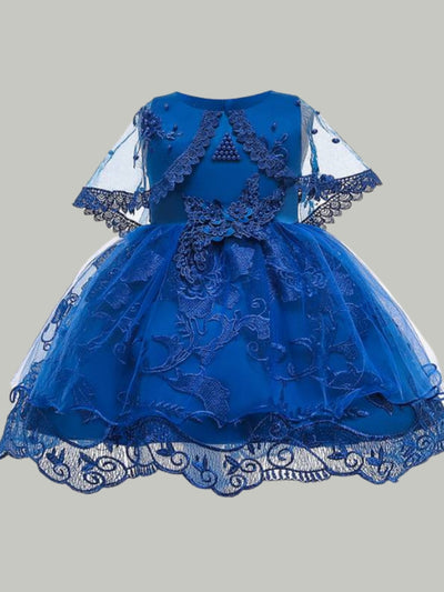Girls satin dress has an embroidered tulle overlay with flower applique and pearl details. The dress has a tulle capelet and a large bow on the back