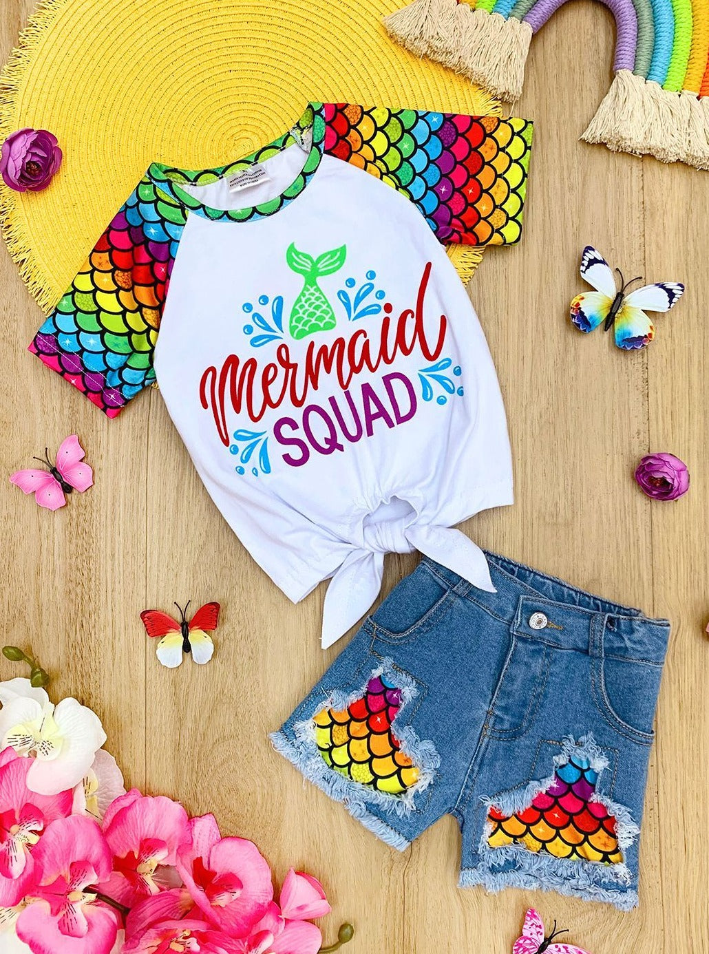 Girls Summer set features a rainbow mermaid scale raglan sleeve knot hem "Mermaid Squad" graphic tee with matching patched distressed denim shorts 2T to 12Y