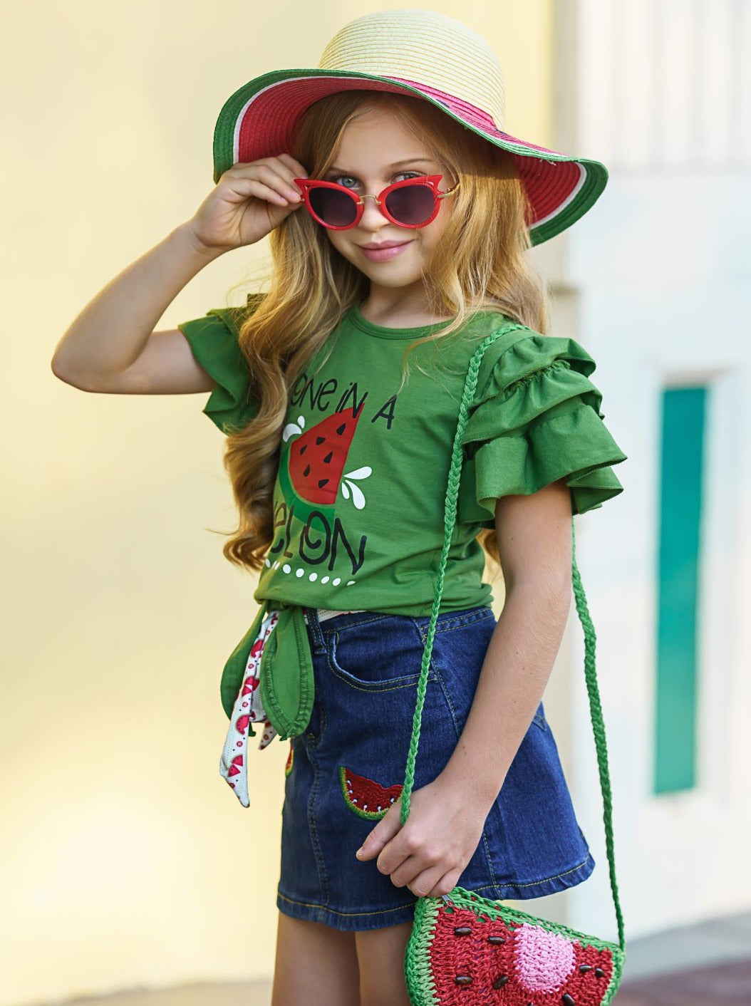 Girls Spring set features a green top with multi-layer ruffled sleeves, "One in a Melon" graphic print, and a denim skirt with watermelon applique and a sash belt