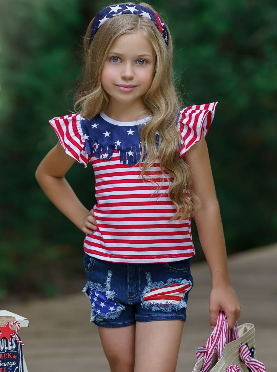 Girls 4th of July set features an American flag print flutter sleeve top with fringe neckline and patched denim shorts with a sash