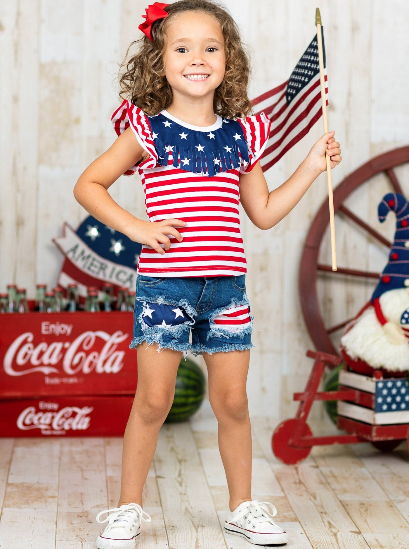 Girls 4th of July set features an American flag print flutter sleeve top with fringe neckline and patched denim shorts with a sash