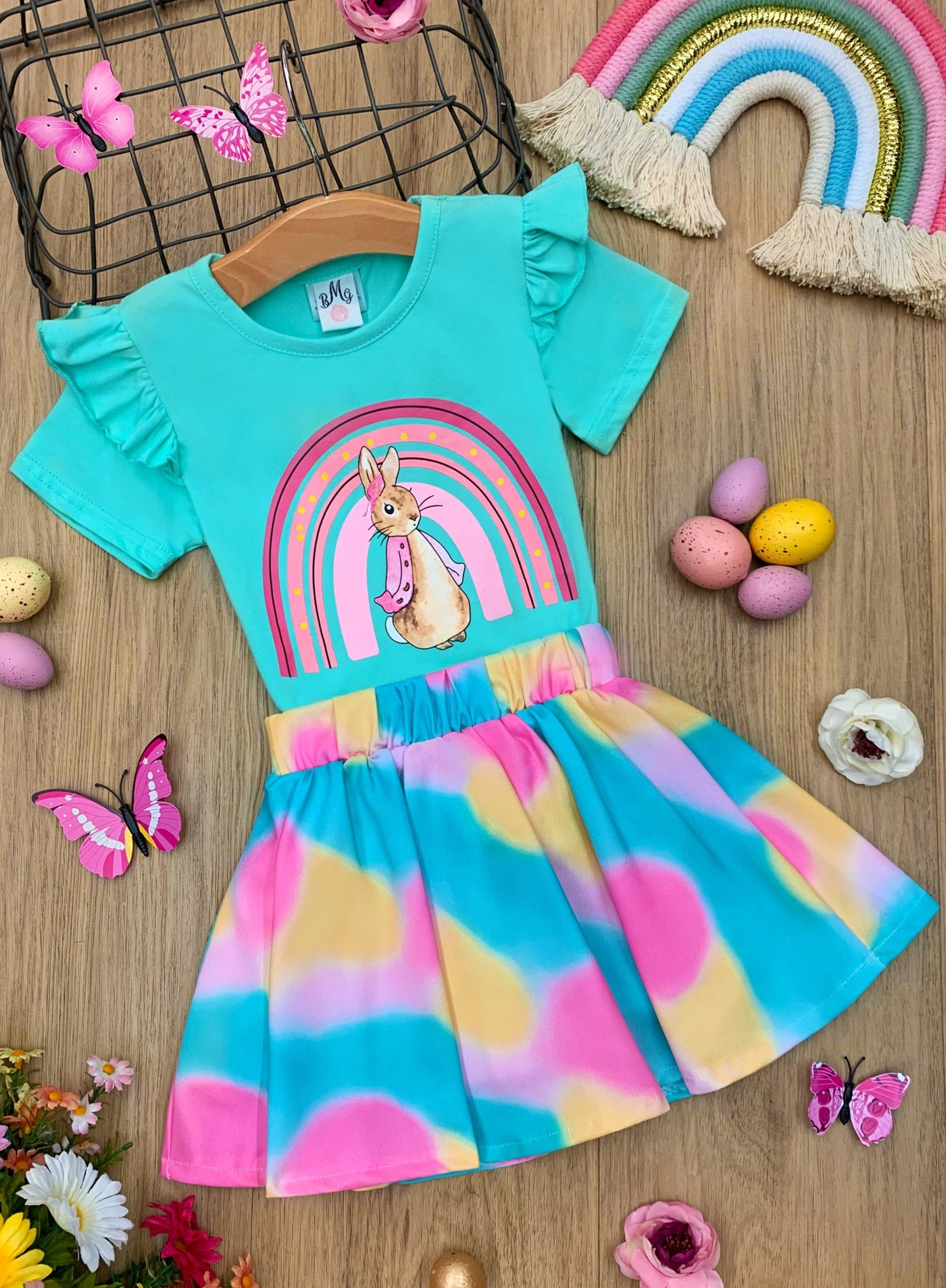Features a mint ruffle shoulder, knot-hem top with a bunny and rainbow graphic design, and a vibrant multicolor skirt for 2T to 10Y toddlers and girls